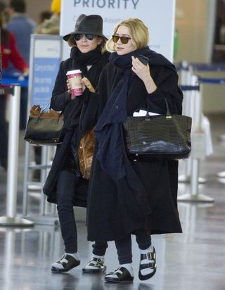 Mary-Kate and Ashley Olsen walking in an airport wearing oversize coats, large scarves, socks with Birkenstock sandals, and The Row bags.