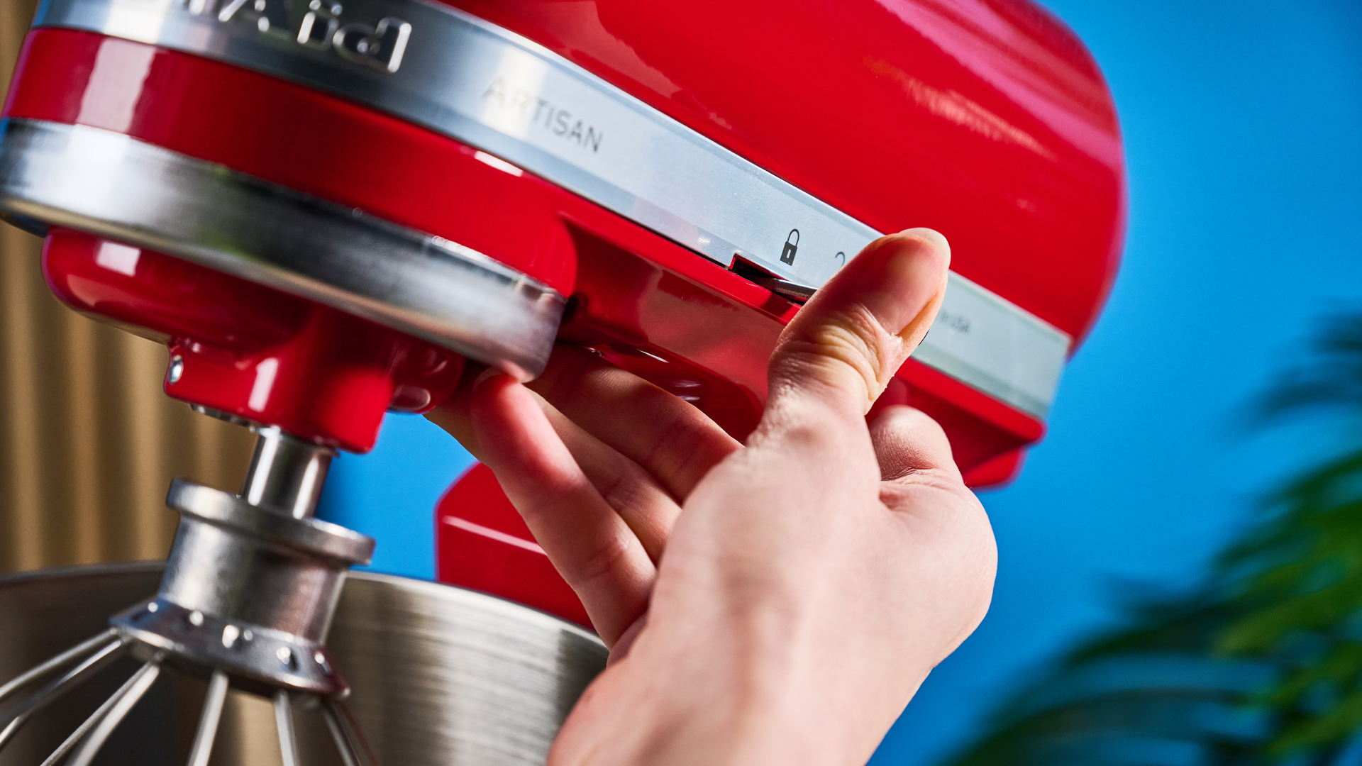 photograph of the kitchenaid artisan stand mixer tilt head in cherry red, with the mixing bowl, paddle, dough hook, and whisk attachments, photographed against a blue background