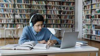 Best laptops for students - Student with long hair is sitting in the library wearing glasses and headphones. Surrounded by books, he's looking at his laptop and writing notes in pencil.