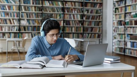 Student with long hair is sitting in the library wearing glasses and headphones. Surrounded by books, he's looking at his laptop and writing notes in pencil.
