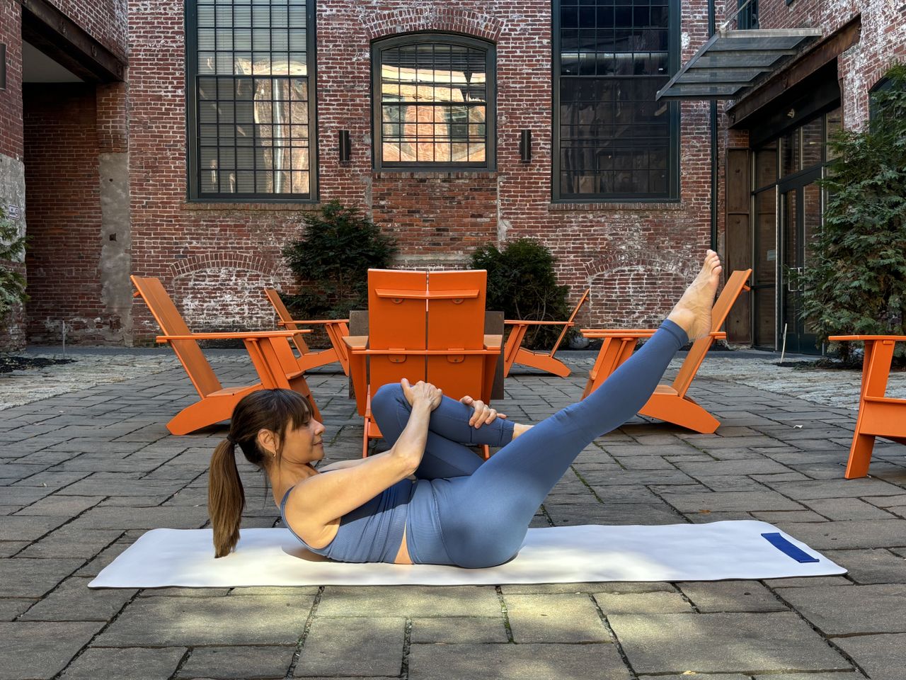 Pilates teacher Monica Borodin performs a single leg stretch. She is in a vest and leggings and exercises on a mat outside. Behind her we see a brick walled building with large windows and a table and chairs. She is lying on her back, holding her left leg close to her chest while her right leg is extended in the air.
