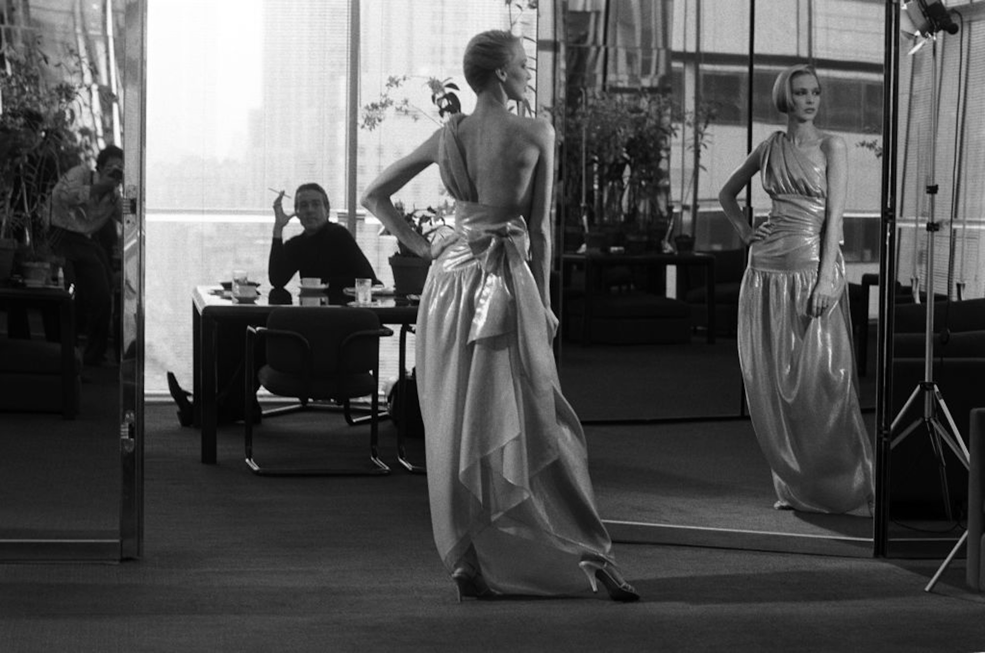 halston at a desk looking on as a model gazes at her reflection