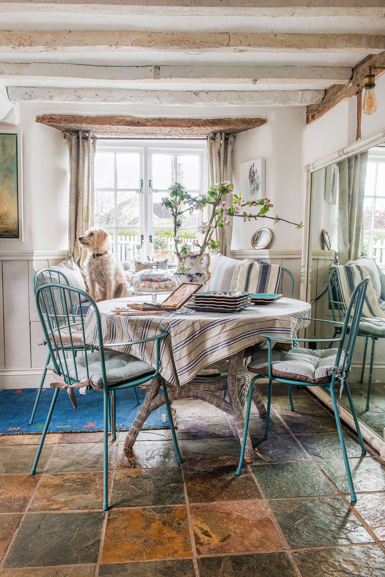 round table in dining room with striped cloth and blue chairs and dog on window seat pale painted walls and whitewashed beams