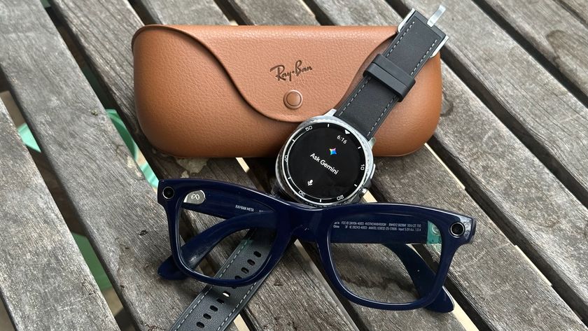 A photo of the Ray-Ban Meta (Gen 2) smart glasses and case sitting on a table with the Samsung Galaxy Watch 8 Classic between them, leaned against the case, with the display showing &quot;Ask Gemini.&quot;