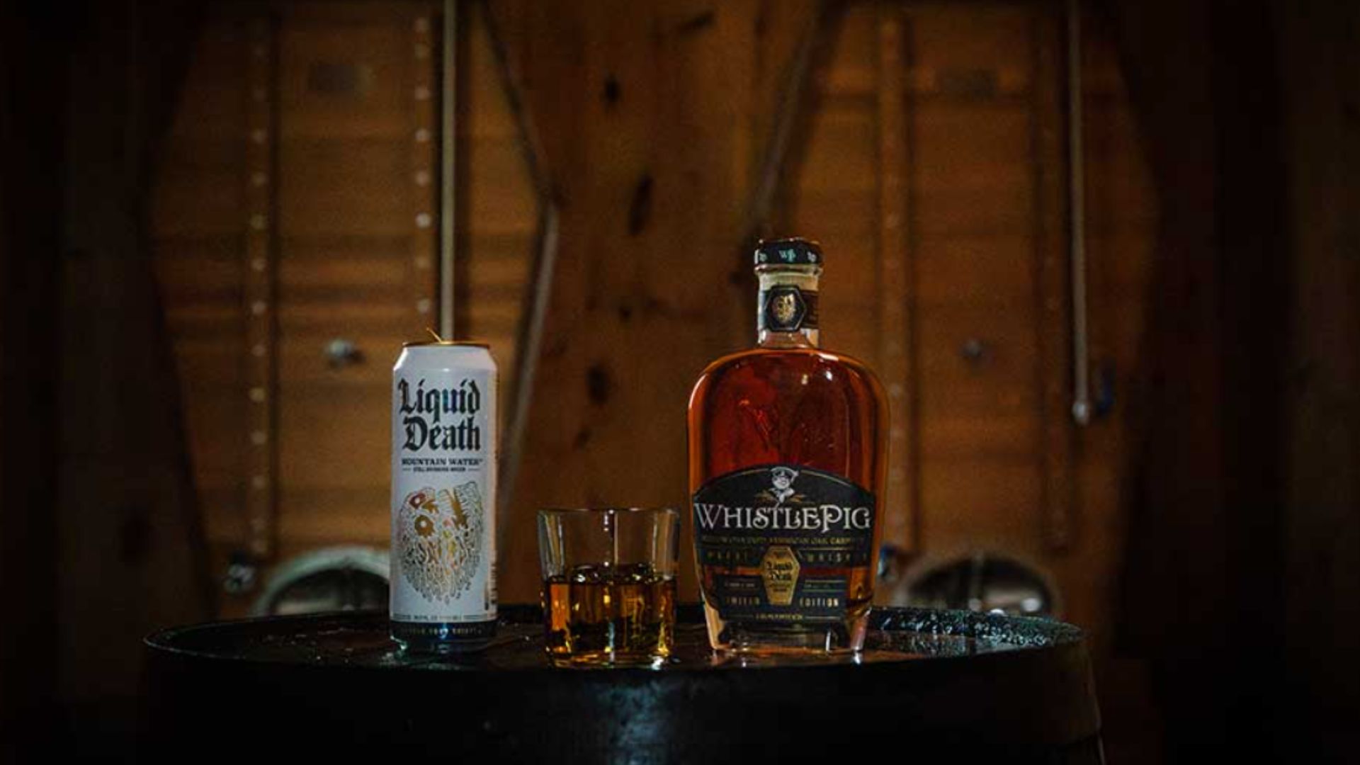 an image of a can of liquid death water on top of a barrel. Next to it is a small tumblr with a double serve of whiskey. On the right is a 750ml bottle of WhistlePig whiskey