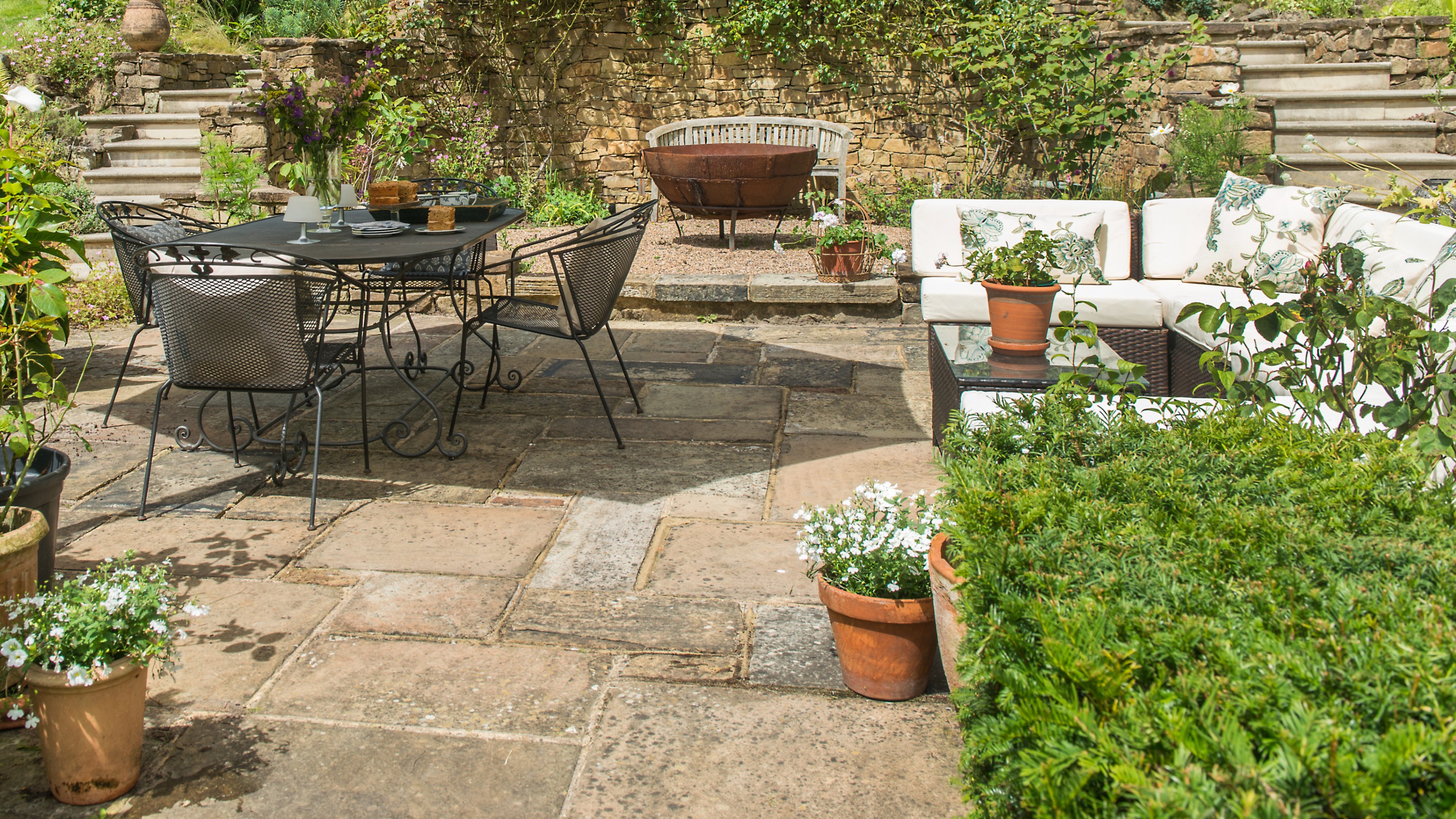 A paved garden with a black metal dining set and a modular synthetic rattan lounge set with cream cushions
