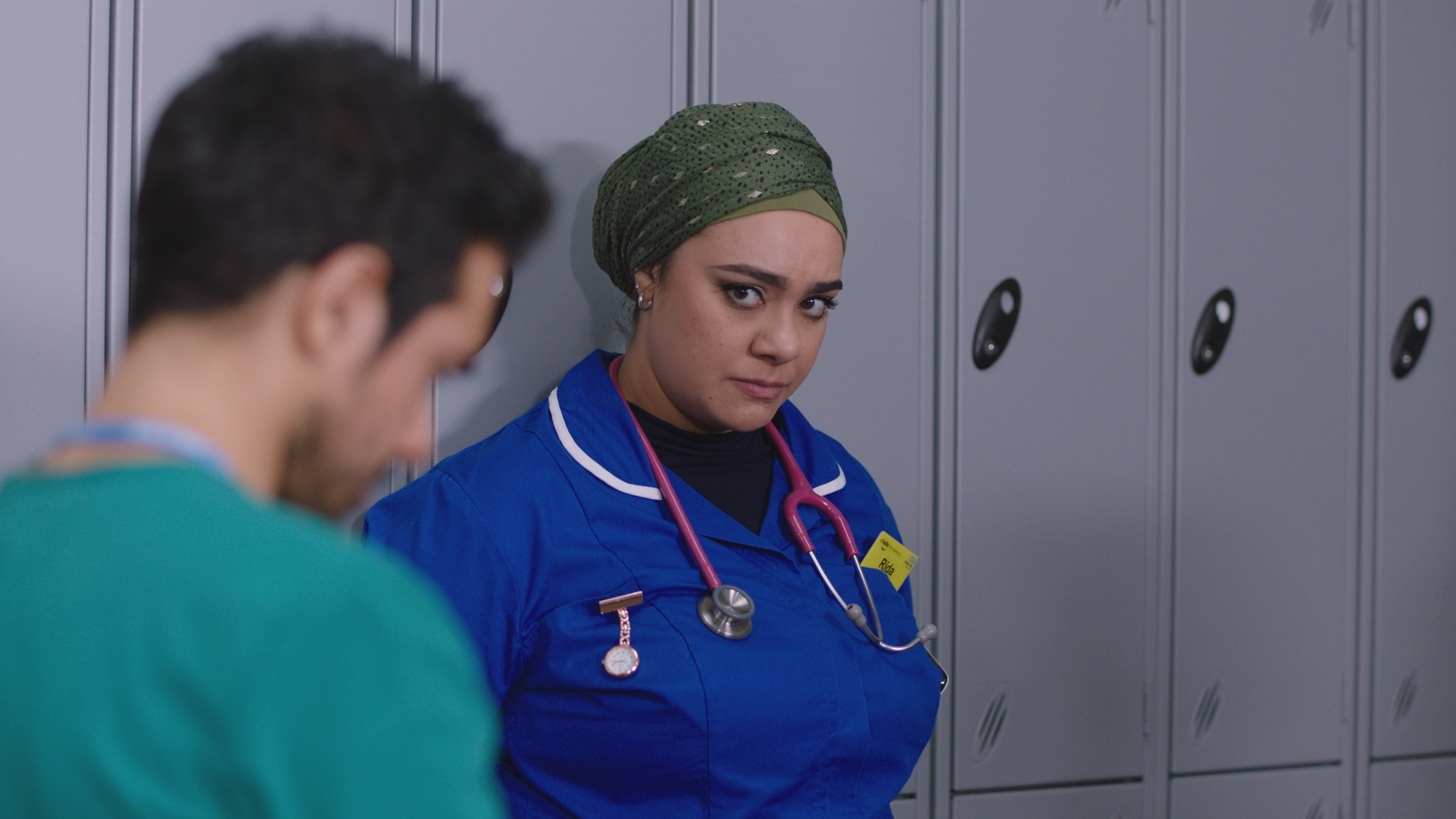 Rida leans against a locker, looking at Rash, as he stares at the ground.
