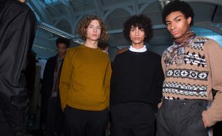 Four male models wearing looks from Margaret Howell's collection. One model is wearing a black coat and dual coloured scarf. Another model is wearing a mustard coloured jumper and black trousers. The model next to him is wearing a white shirt, black jumper and black trousers. And the fourth model is wearing a brown jumper with black, white and grey pattern, a dual coloured scarf tied around his neck and grey trousers