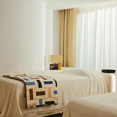 A minimalist bedroom with the bed covered in a white sheet with the Zara Home x Collagerie quilt on top and table lamp on the bedside table