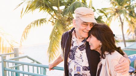 Older couple hugging, with a palm tree and the ocean in the background