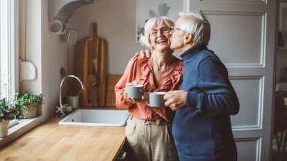 An older couple laugh and smile as the man kisses the woman on the cheek in their kitchen.