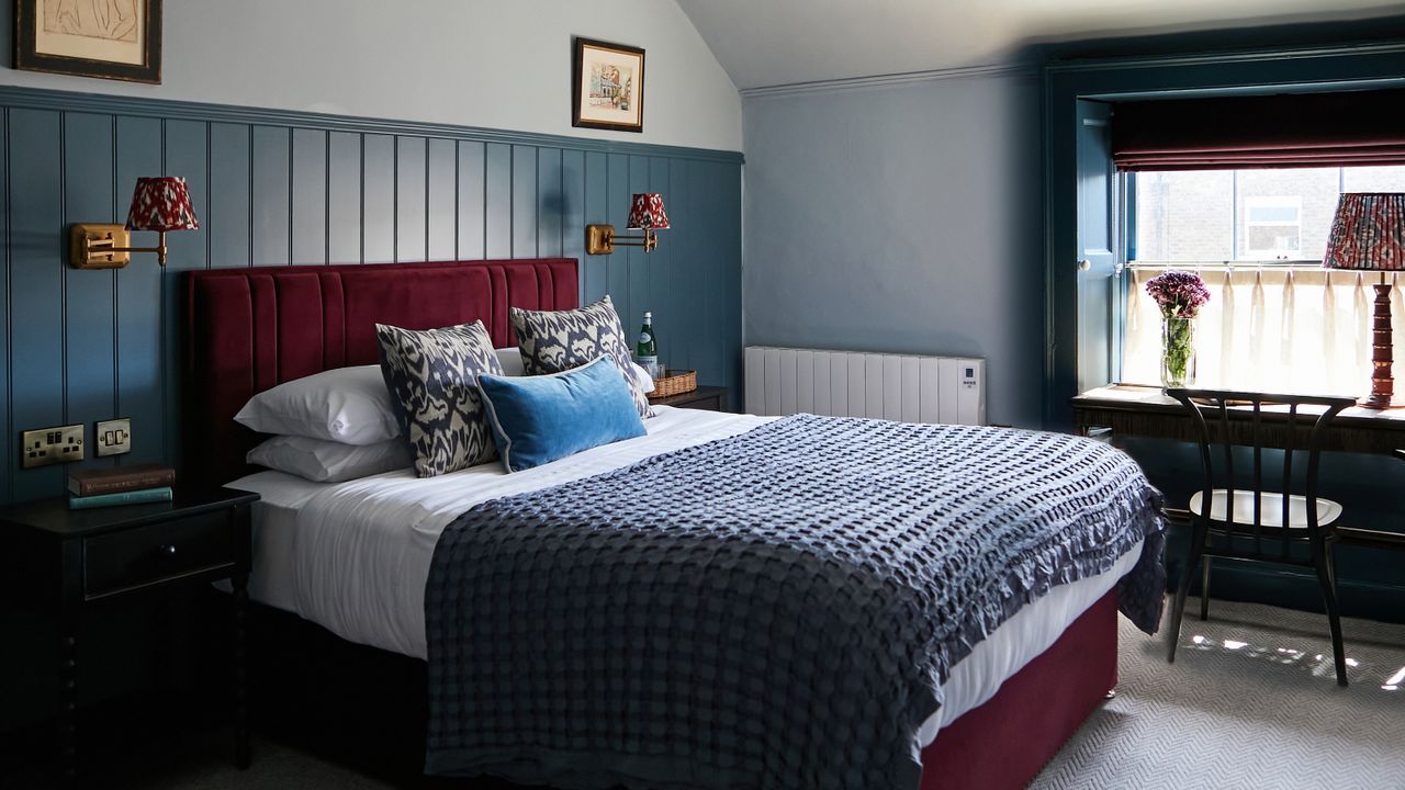 bedroom with blue half panelled wall, red upholstered bed, window with blind and chair in front
