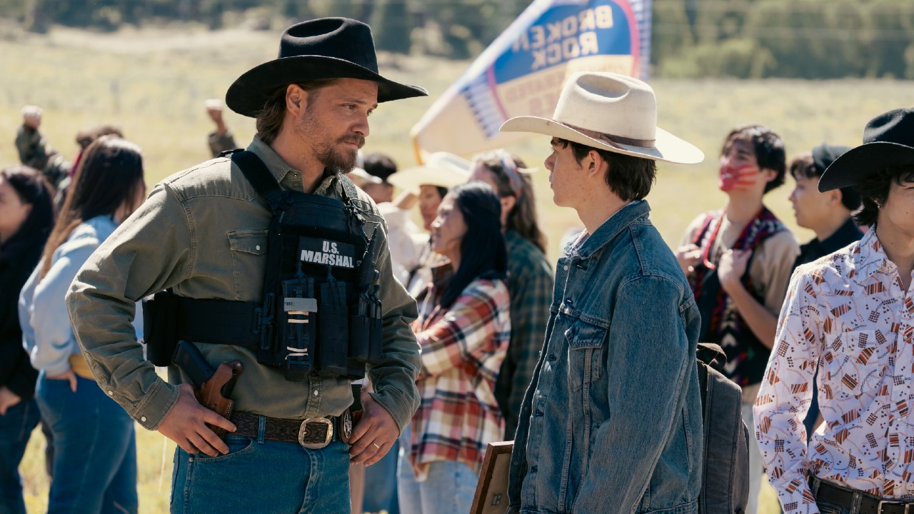 (L-R): Luke Grimes as Kayce Dutton and Brecken Merrill as Tate Dutton standing next to each other in front of a group of people. 