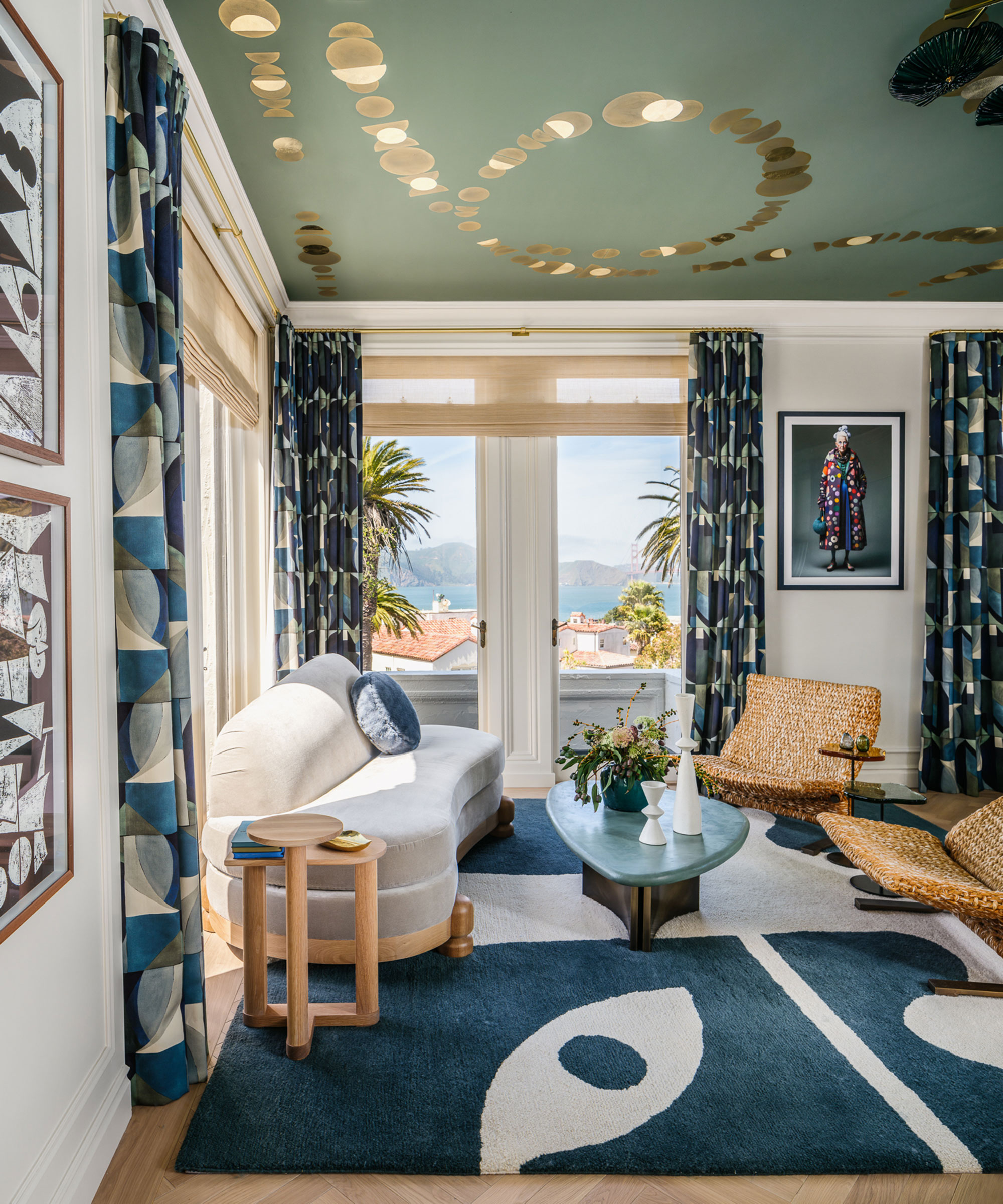 A chic living room with a sage green ceiling featuring gold moon-phase patterns. It includes a curved sofa, woven chairs, and a bold blue-and-white rug overlooking a sunny coastal view.