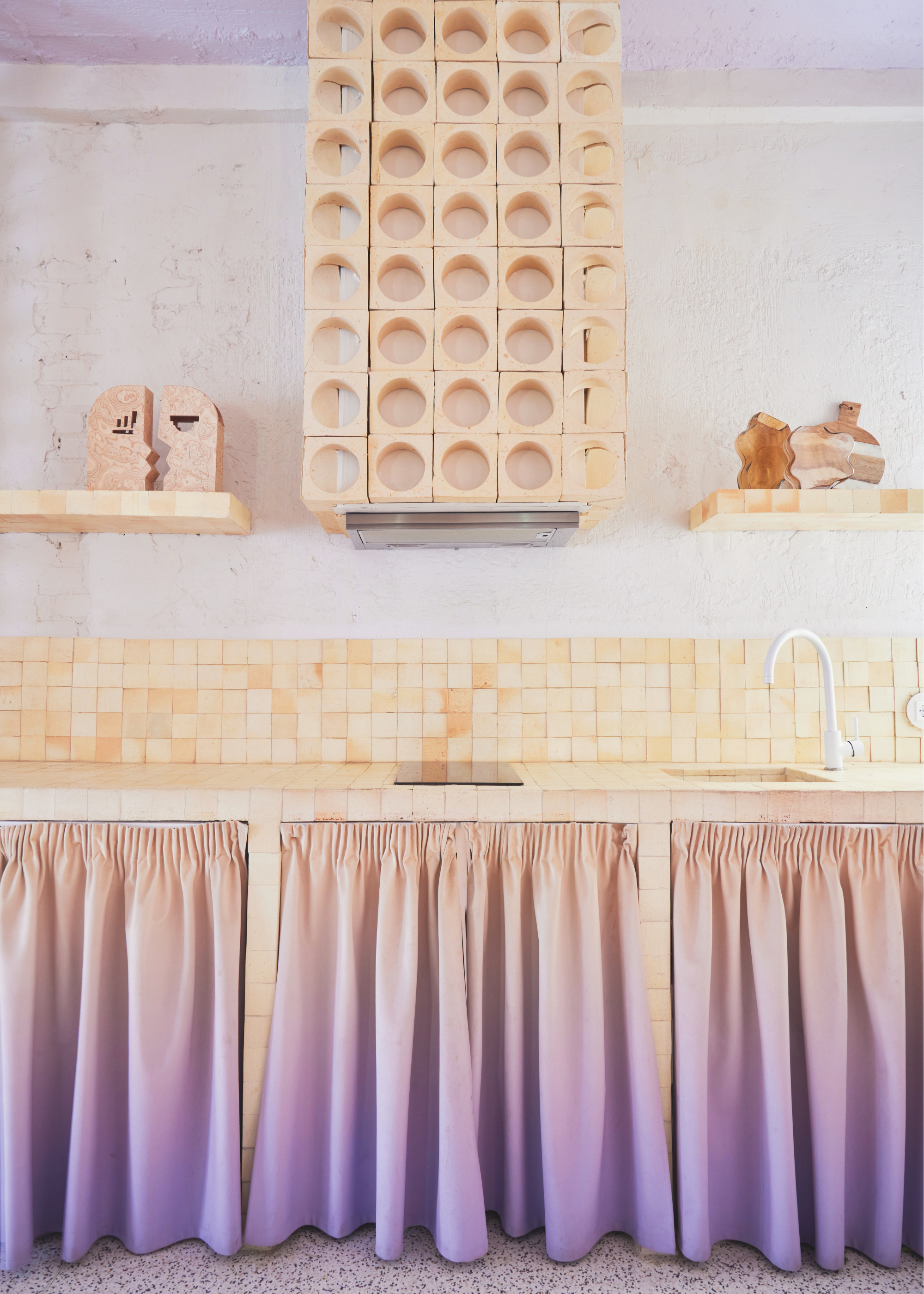 a cream and purple kitchen with modern accents including a breeze block extractor hood against a white painted wall and ombre purple curtains in place of cabinets