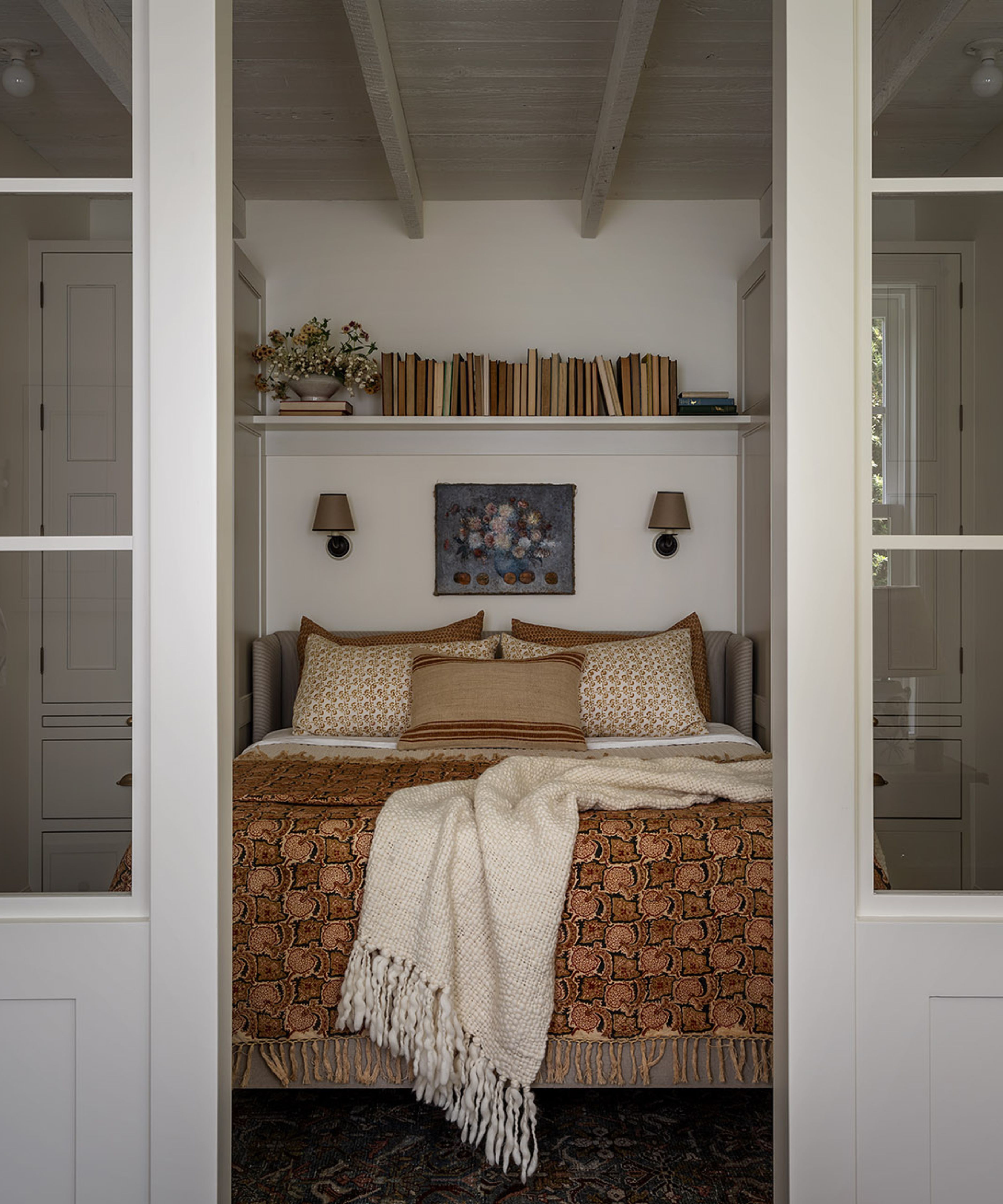 a view through glass partition doors into a white bedroom space with mustard accessories and a small book-filled shelf, art, and two sconces hung above the bed