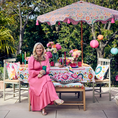 Sophie Robinson in pink dress sat at bench decorated with colourful candles, tablecloth and flowers in a patio garden.