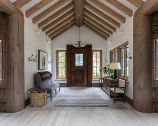 Traditional interior with vaulted ceiling with wooden beams in entryway, looking onto large wooden front door with window either side, small window in door, wooden flooring, bench