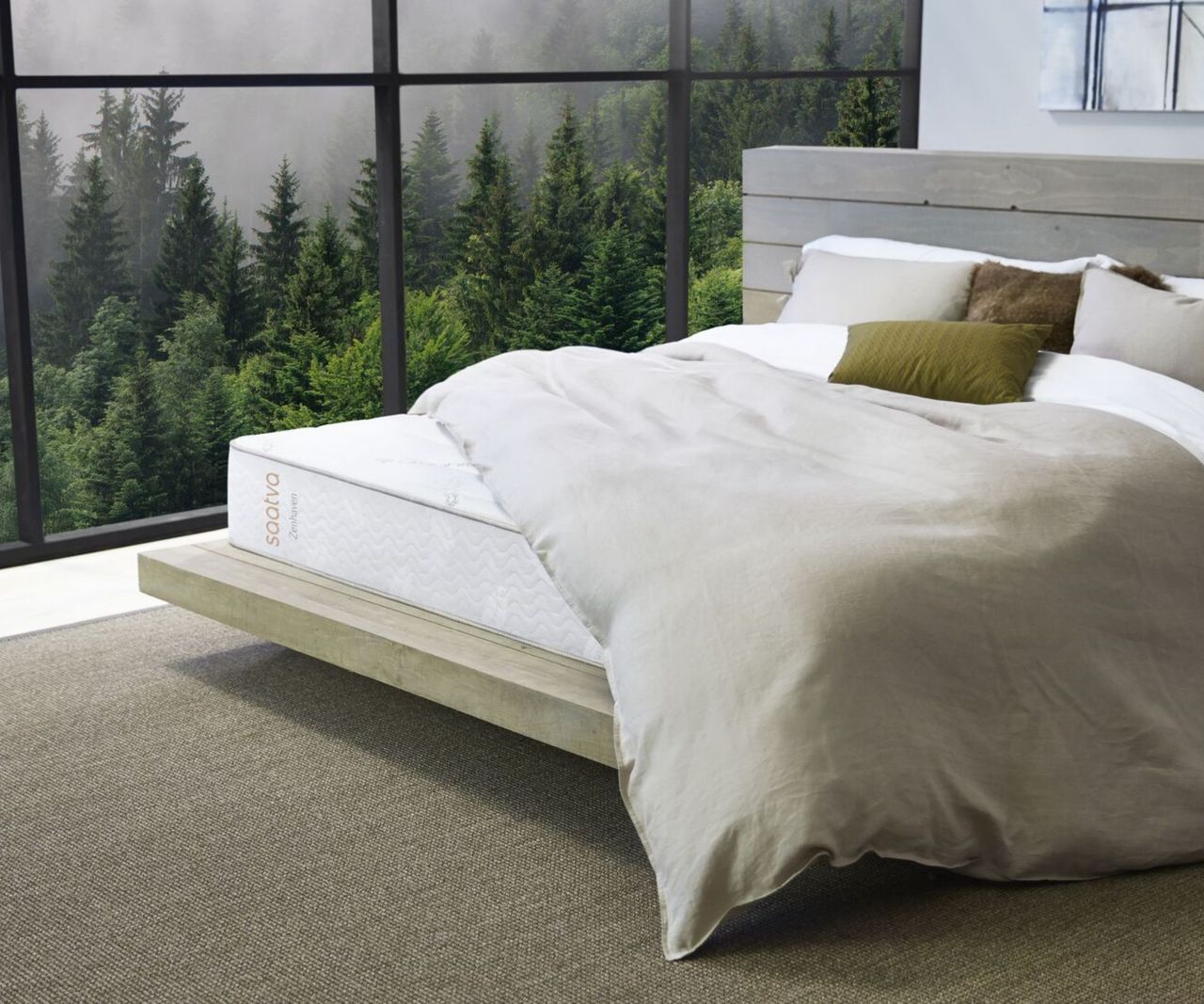 Saatva Zenhaven Natural Latex Mattress on a wooden bed beside a window with a forest view. The bed has neutral beige bedding with green cushions on it, and beneath the bed is a large rug.