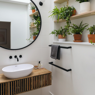 Circle mirro over sink with wooden cabinet. Black towl rail and two shelves filled with houseplants