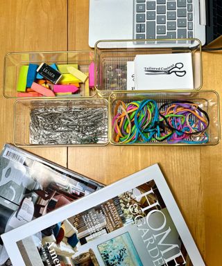 Clear plastic drawer organizers with a gold rim being used to organize small stationary on a wooden desk, beside a Homes & Gardens magazine