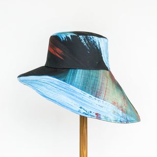 A colorful hat in the tones of black, light blue, sea green, and orange sits on top of a wooden stick against a plain grey background.