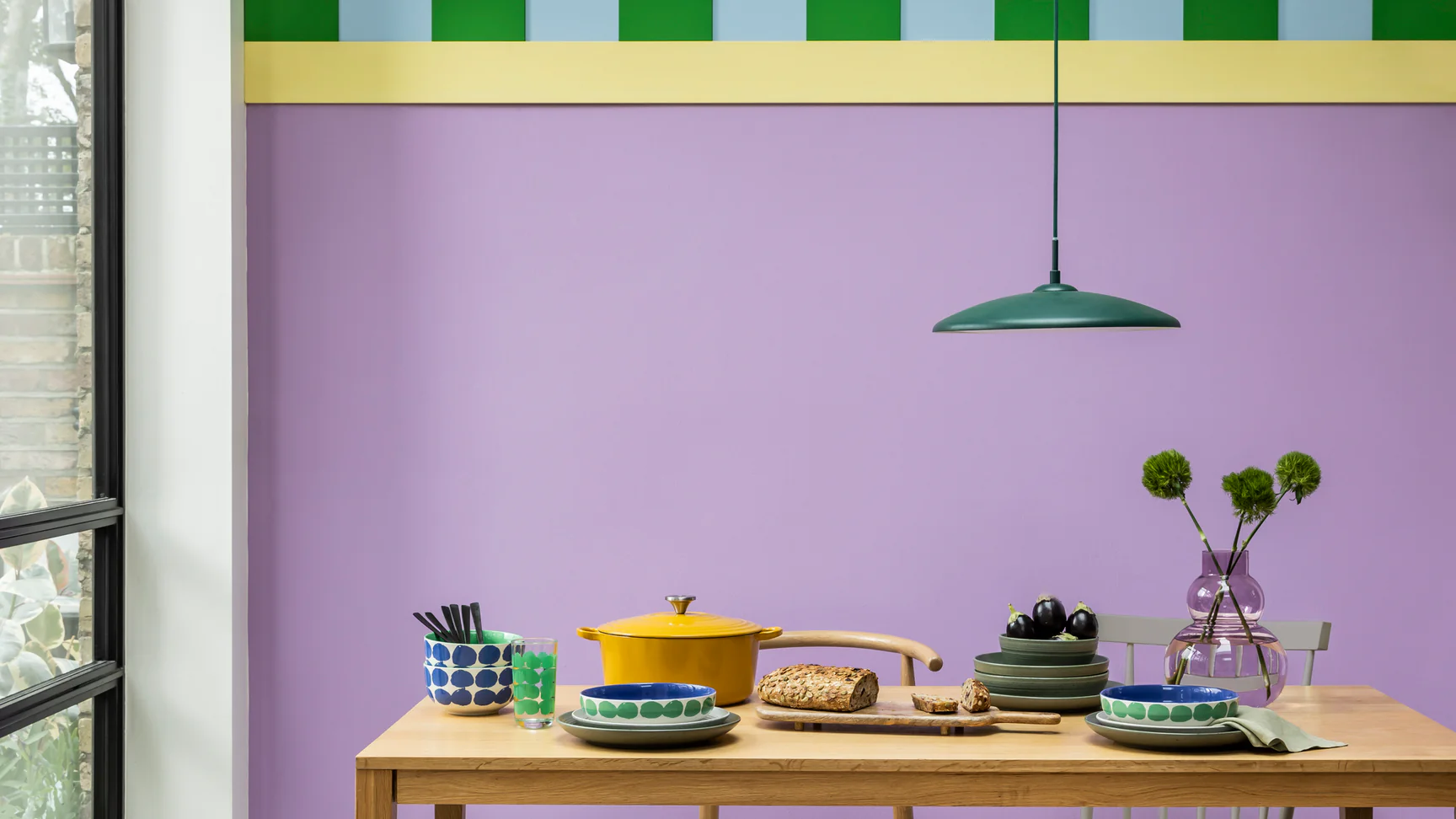 dining room with bright purple wall, yellow trim with a striped blue and green feature, a timber dining table styled with colorful pots and pans and a purple glass vase with flowers