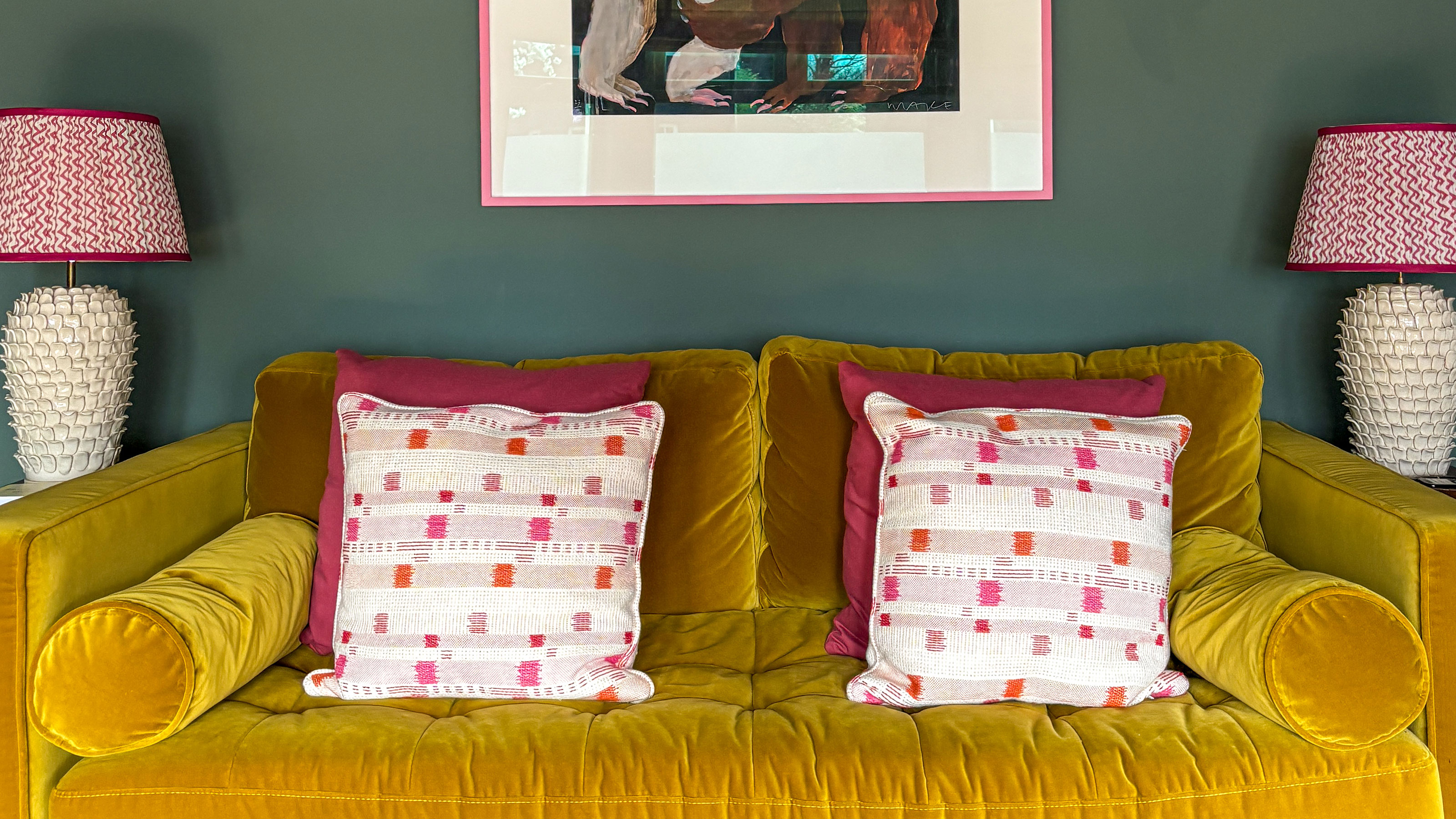 Green living room with mustard-gold sofa with red and white cushions, below a painting and with two table lamps on either side