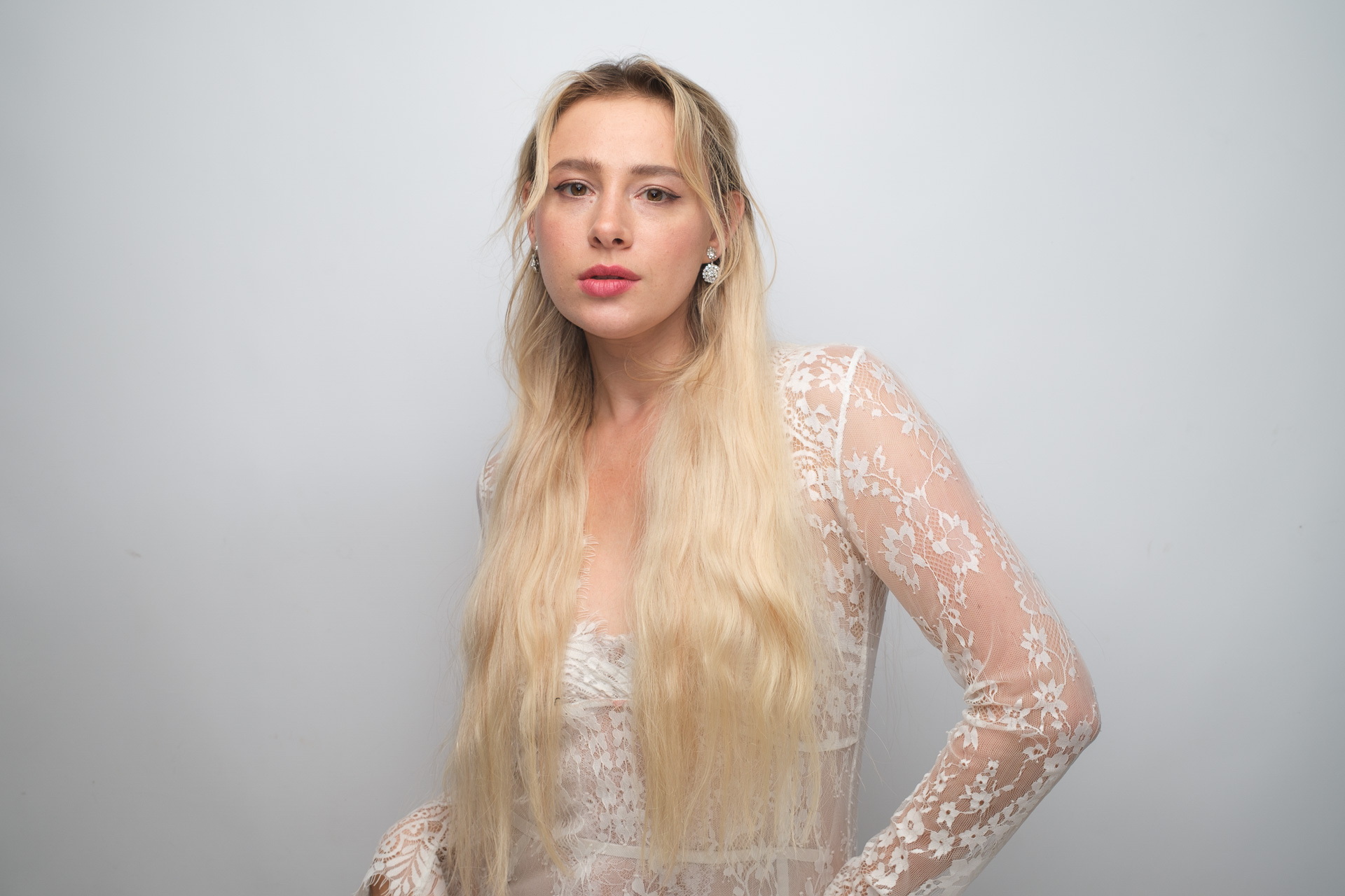 Portrait of a blonde woman in a white lace outfit posing with one hand on her hip against a light-gray background.