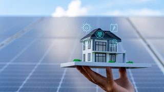 hand holding 3D model of house with symbols around it and solar panels in the background