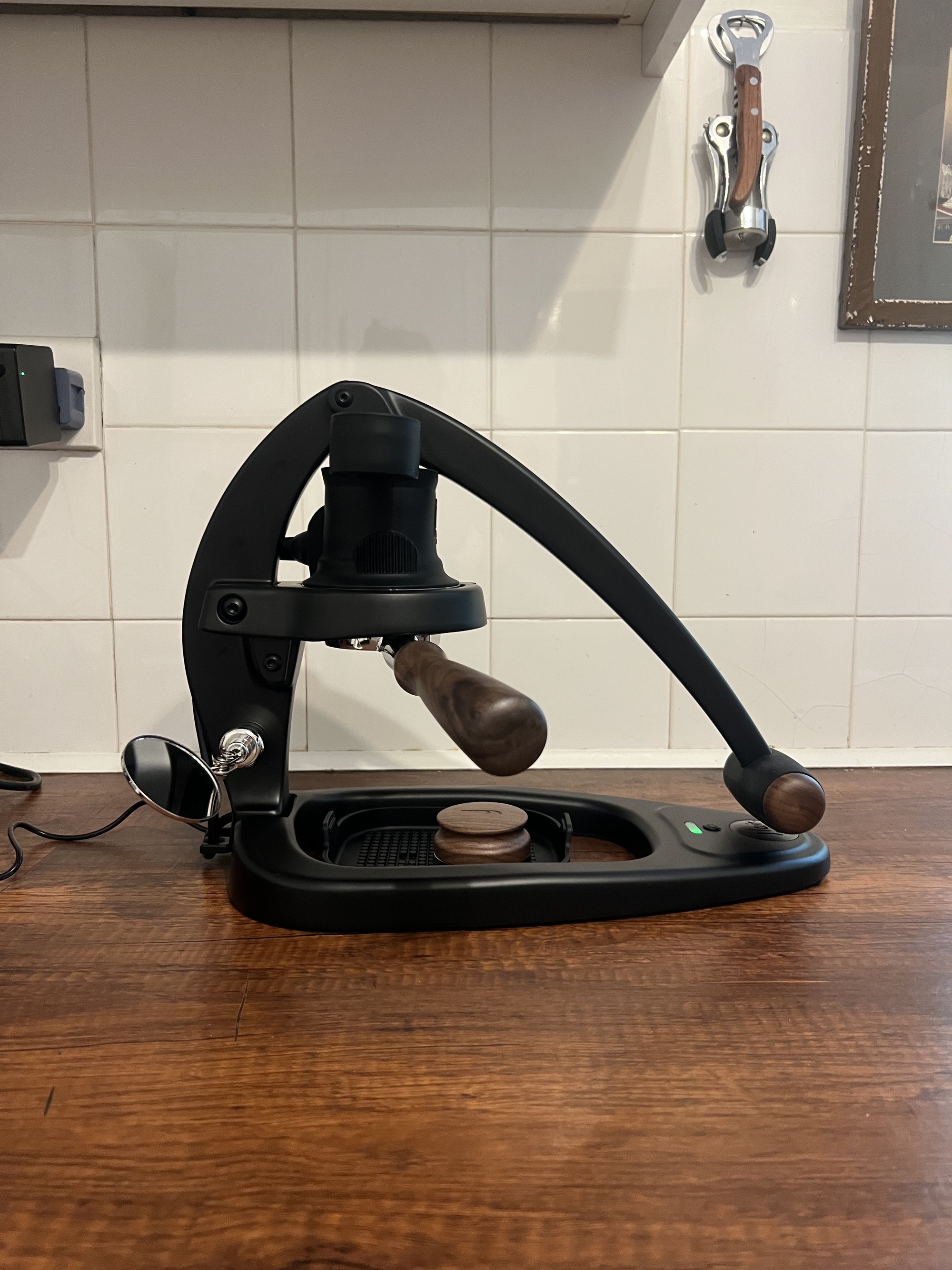 Image of a black and dark wood manual espresso maker on a wooden countertop in a kitchen with a white tile backsplash.