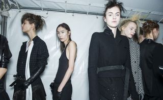 Three female models, one wearing a blue plaid shirt with white silk detailing, one in a black vest and trousers, and one in a black jacket