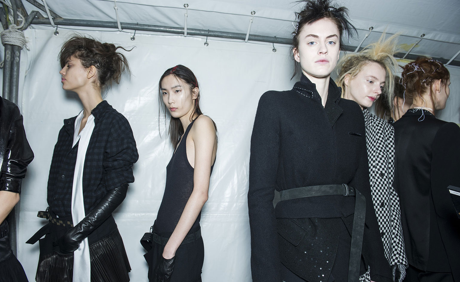 Three female models, one wearing a blue plaid shirt with white silk detailing, one in a black vest and trousers, and one in a black jacket