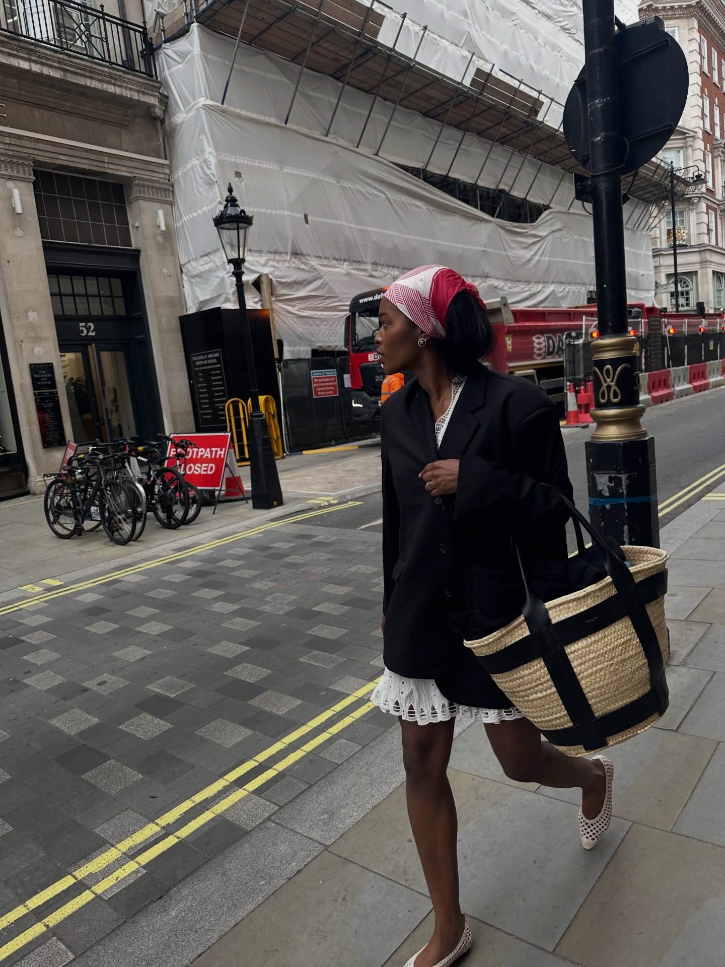 Fashion person wears AnneMarlin June Outfit Ideas 2026. She wears a black blazer over a white Broderie Anglais dress, knitted flats and carries a DeMellier Sardinia bag.