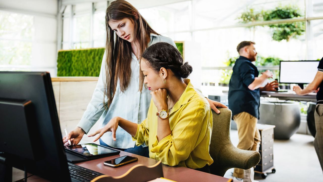 An employee stands at the desk of another employee while they look at a tablet together.