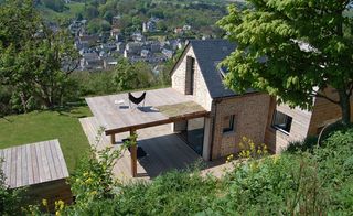 Daytime, outside high view of VW House by Franklin Azzi, brick house, grey tiled roof, windows, wooden balcony, grass lawn, trees, view of surrounding buildings and landscape in the distance