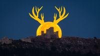 A yellow-orange full moon with antler-like horns rises behind a castle on a hillside in a dark sky.