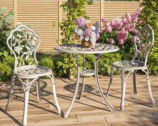 White ornate metal bistro patio table with matching chairs on a wooden decking with vase of summer hydrangea blooms in white purple pink orange on table