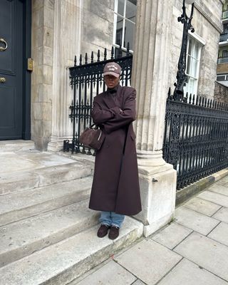 woman wearing long brown coat baseball hat and loafers