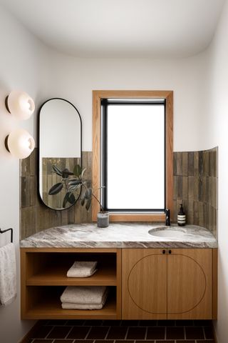wood bathroom vanity with two shelves and gray marble worktop, dark green tiles, large window and oval mirror
