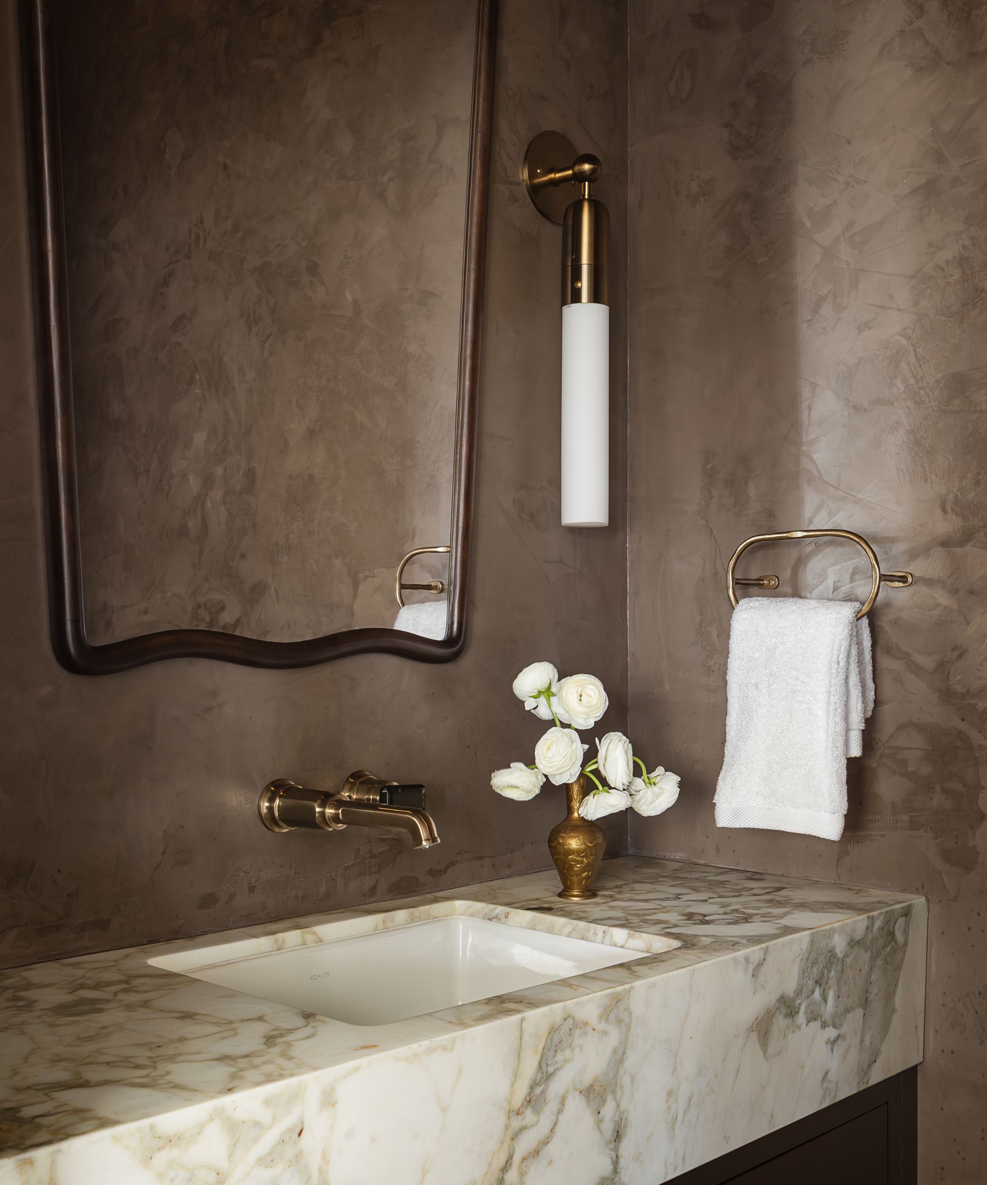 a deep brown plaster finished powder room with marble sink and a wavy brass mirror