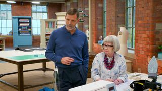Patrick Grant and Esme Young standing at a desk and watching someone sew on the Sewing Bee set 