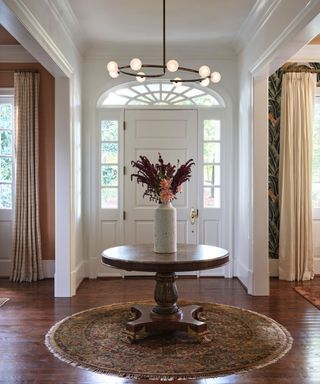 A white entryway with a sculptural light fixture, a circular rug, a circular wooden table, and a vase of flowers