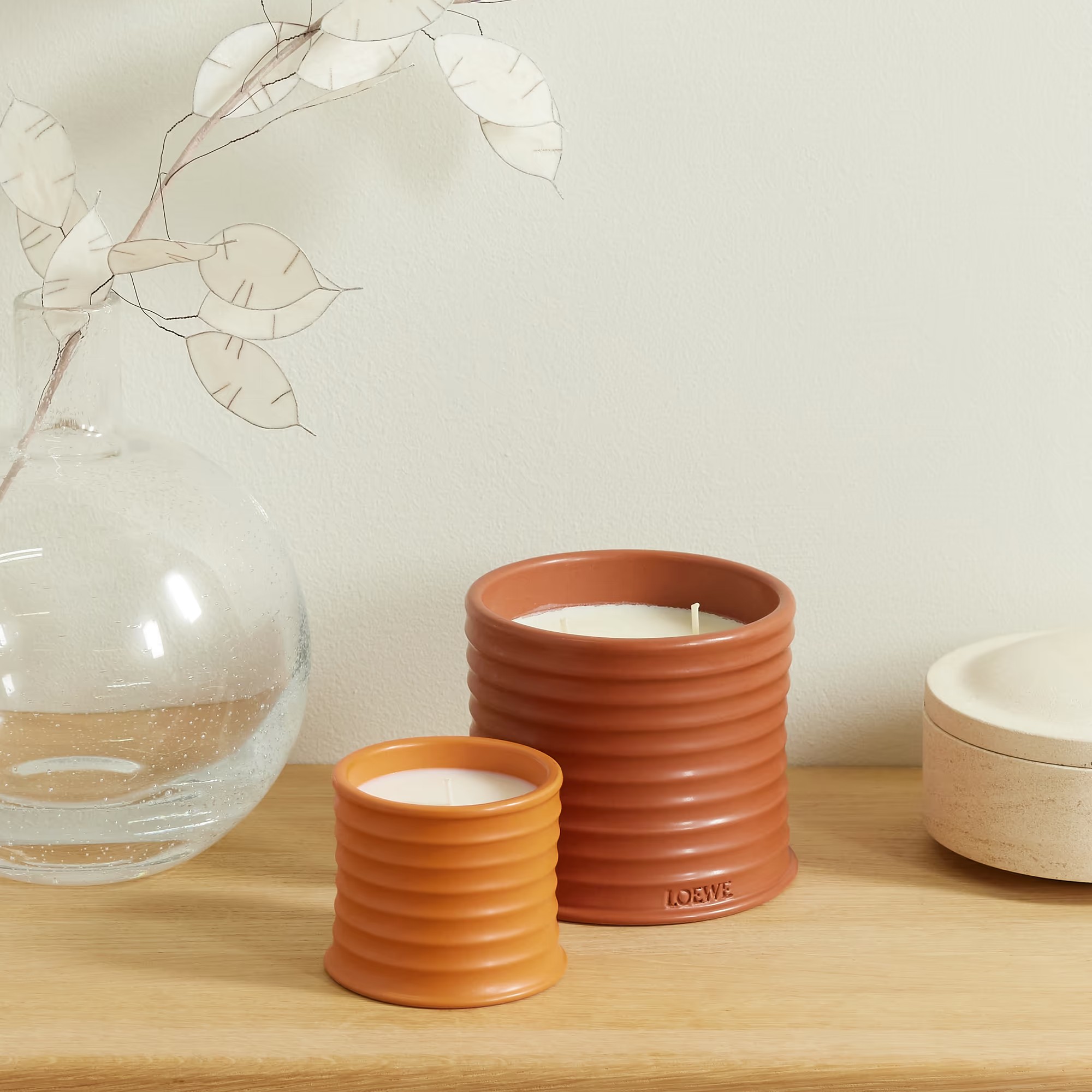 Two orange Loewe scented candles in two different sizes on a wooden sideboard with a round glass vase