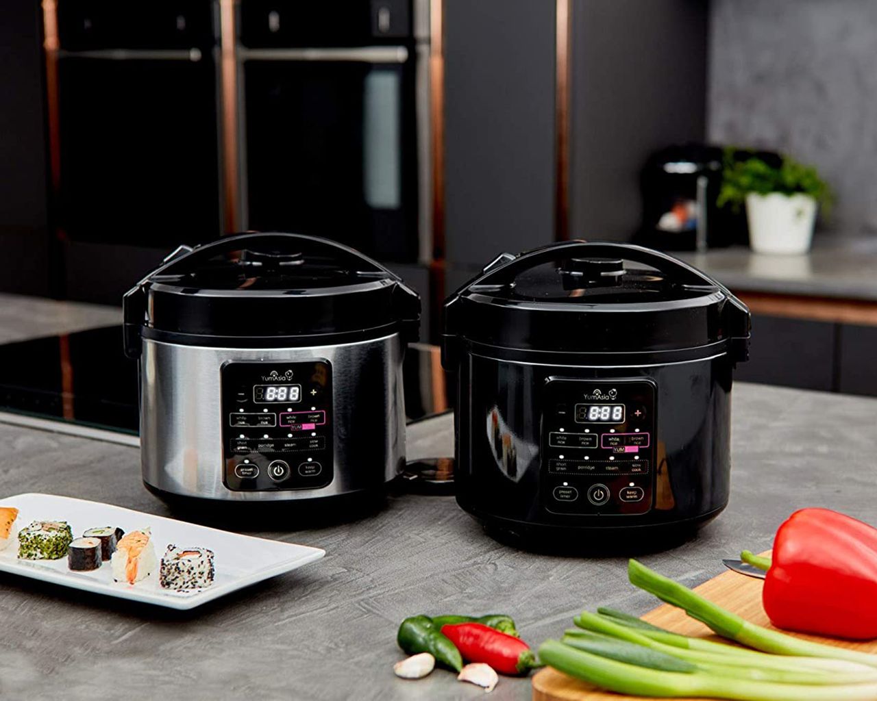 Yumo rice cooker appliances in dark stainless steel and black on worktop in kitchen with plate of sushi, bell peppers, chilli and garlic
