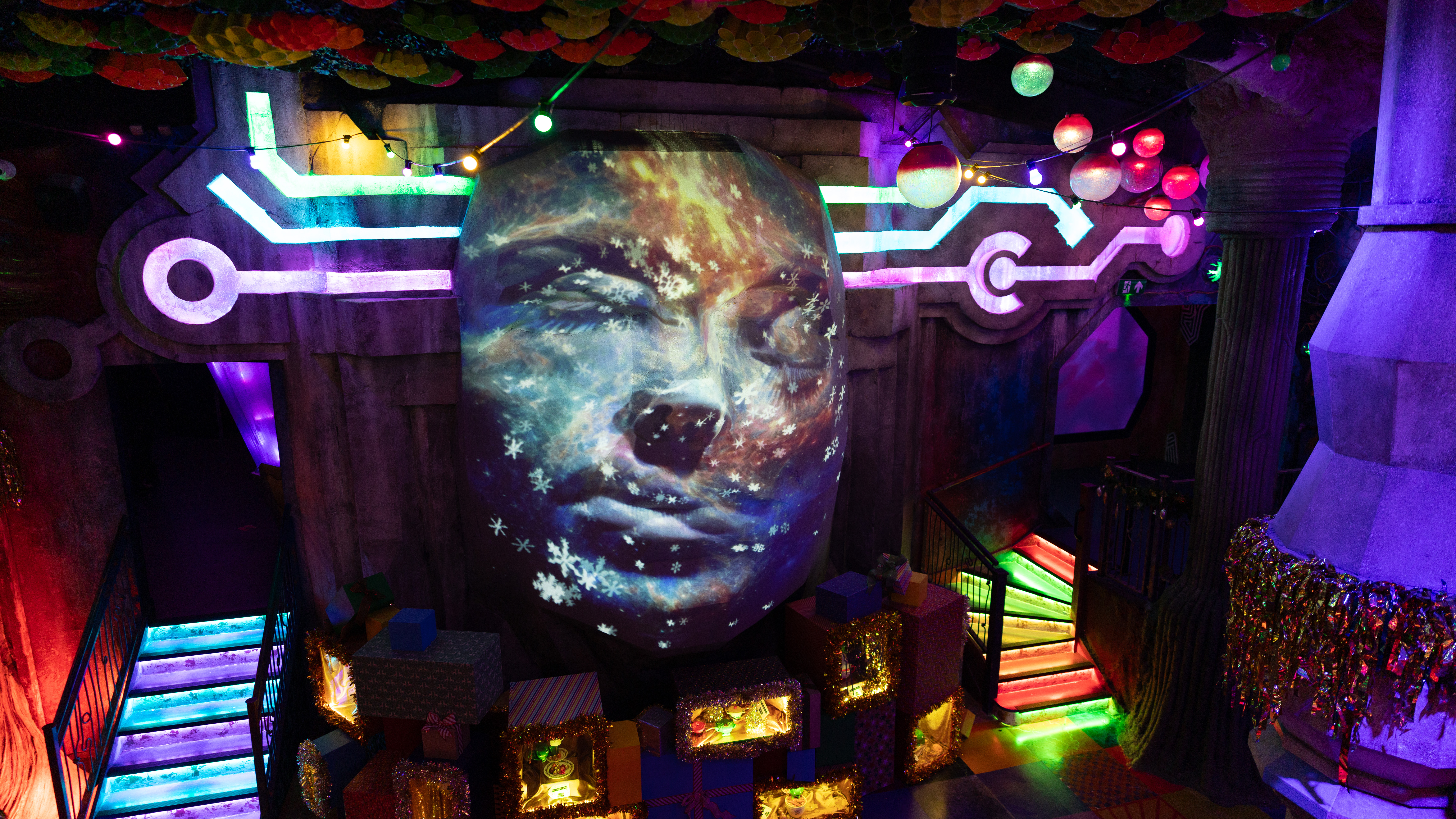 Scene from an indoor theme park, where a large face is being projected with distortion onto a fluorescent backdrop.