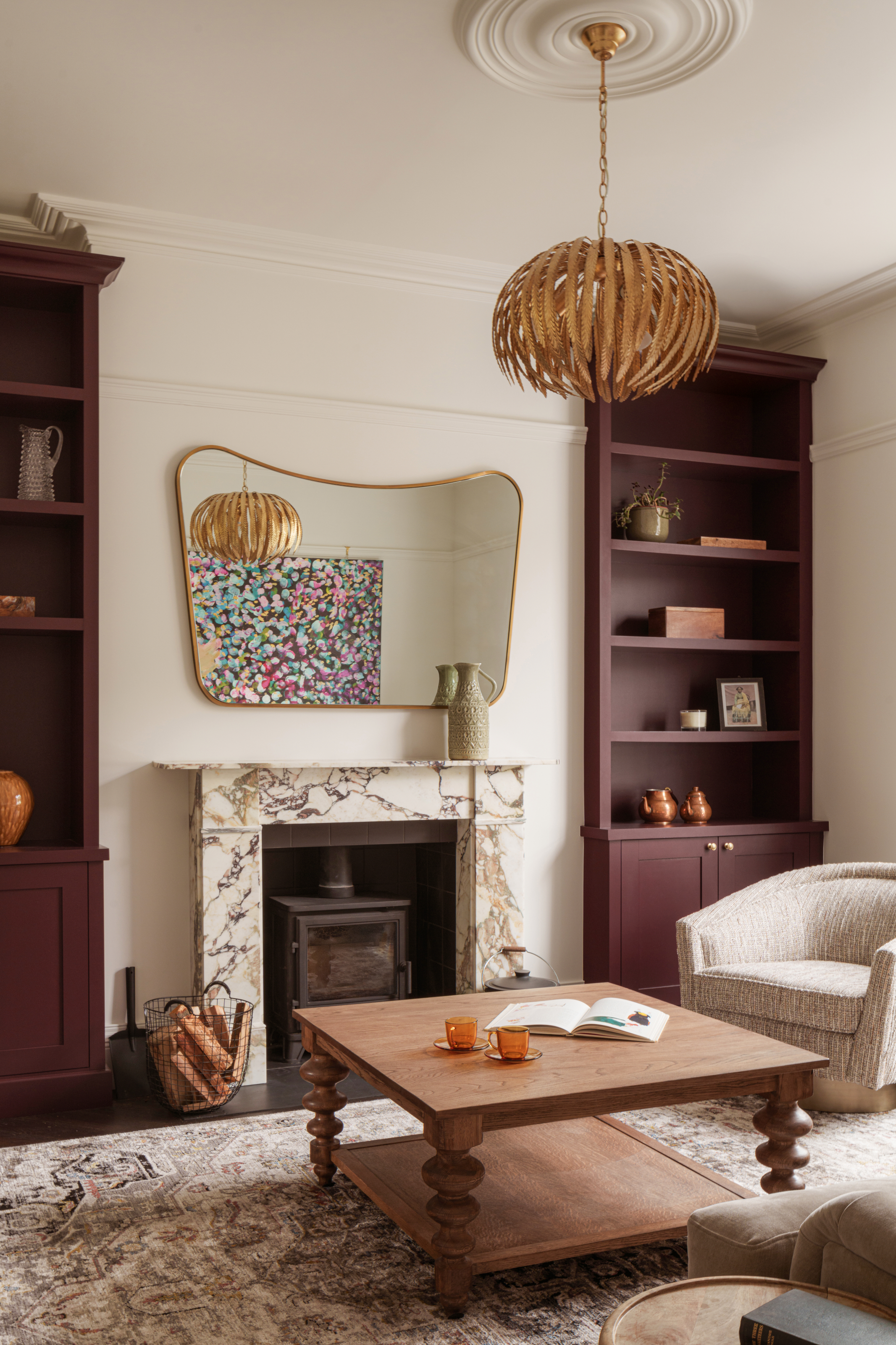 Cream living room with burgundy painted shelves either side of alcove, wood coffee table, marble fireplace and brass mirror