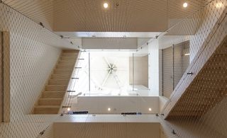 Interior view from below of the shaft at the Super Cube through mesh. The space features white walls, wooden stairs and spotlights