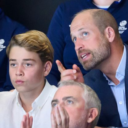 Prince George looks serious and wears a white shirt while his dad Prince William wears a navy suit jacket and points at something, he has a beard but no hair