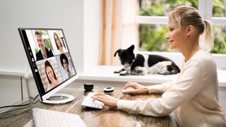 A woman sitting at a desk at home speaking on a video call with her small black and white dog sitting on a windowsill watching her